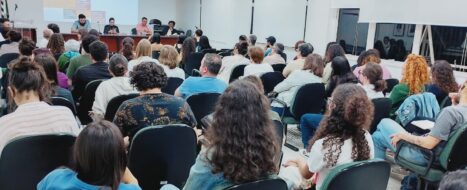 Imagem em plano aberto da plateia do X Fórum, com mesa de debates ao fundo. Comunicação e Trabalho